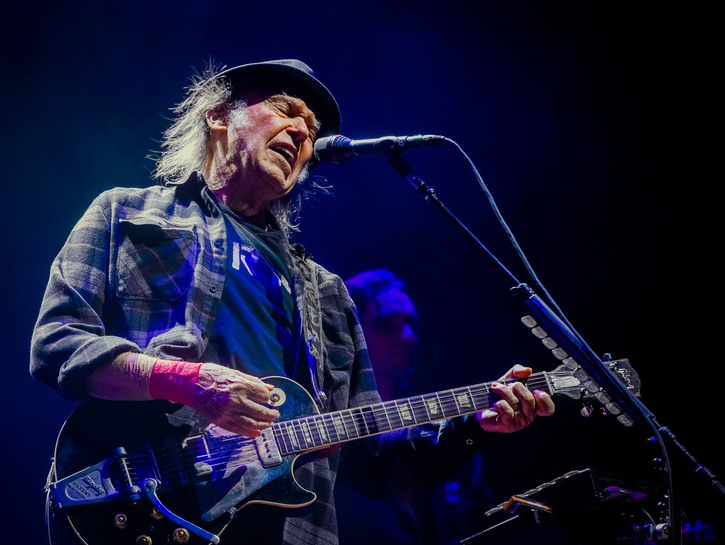 Neil Young playing guitar on stage in a blue flannel shirt and hat in 2019.