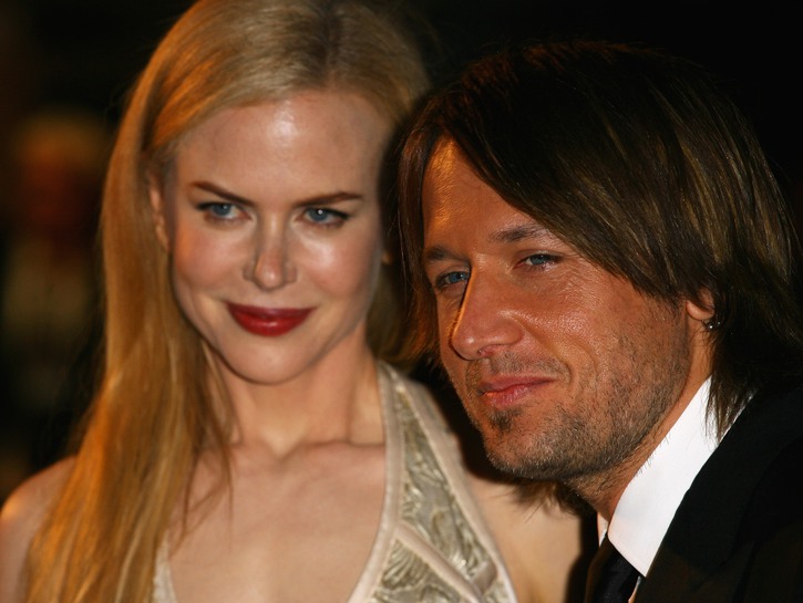 Nicole wearing a gold dress and bright red lipstick stands with husband Keith Urban, in a black suit