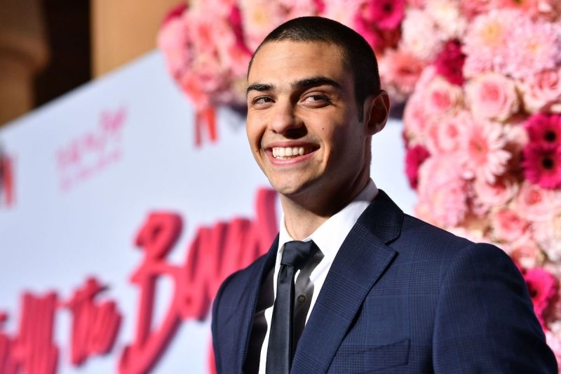Noah Centineo smiles at the camera with a buzzed haircut in a navy blue suit with white shirt