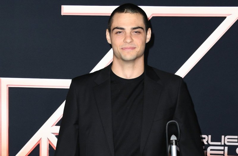Noah Centineo smiles in a black suit with black shirt with a visible scar on his hairline