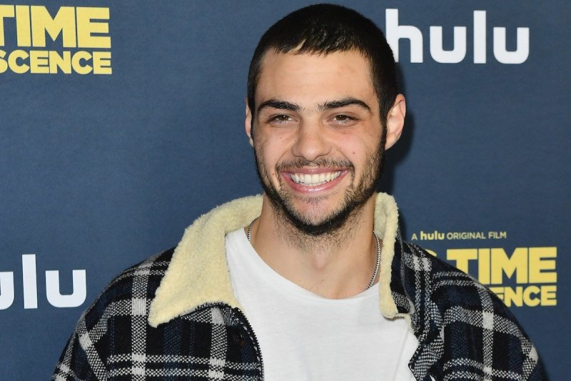 Noah Centineo smiles to his right in a white shirt and plaid sherpa jacket