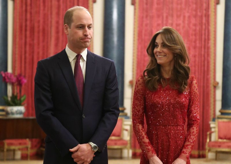 Prince William in a black suit standing with Kate Middleton, in a red dress, in Buckingham Palace
