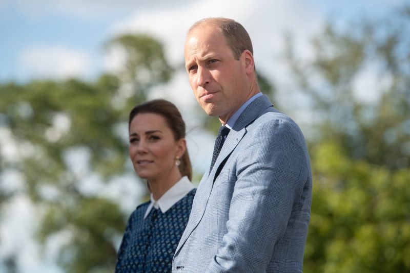 Prince William in a grey suit looks to his left standing in front of Kate Middleton in a blue patter