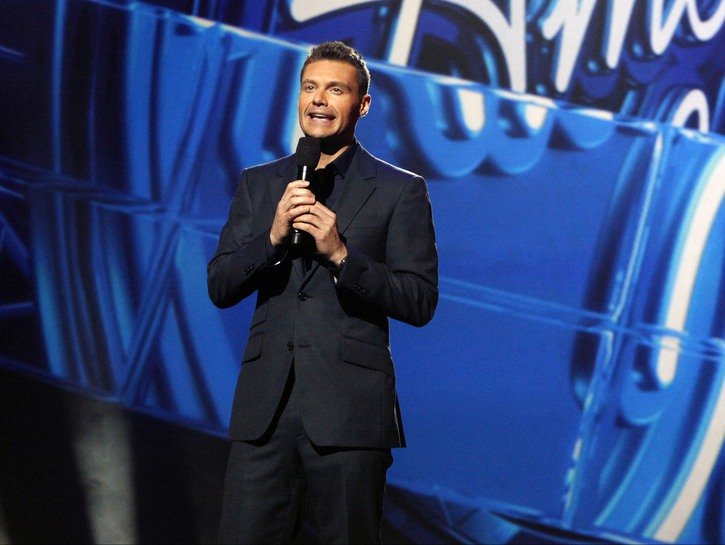 Ryan Seacrest wearing a dark suit onstage at an American Idol event