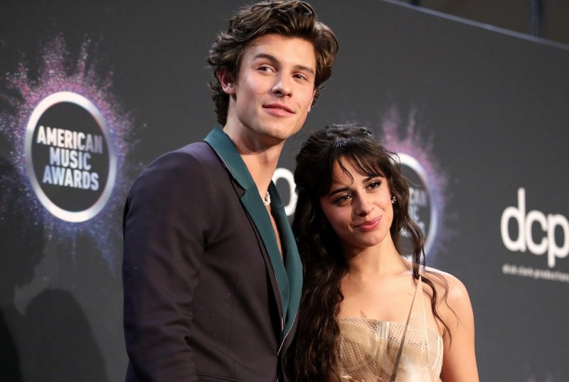 Shawn Mendes in a black and green suit with his arm around Camila Cabello in a tan dress