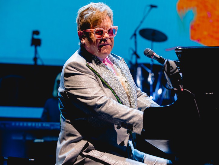 Sir Elton John in a gray suit and pink glasses, performing on stage at his piano.