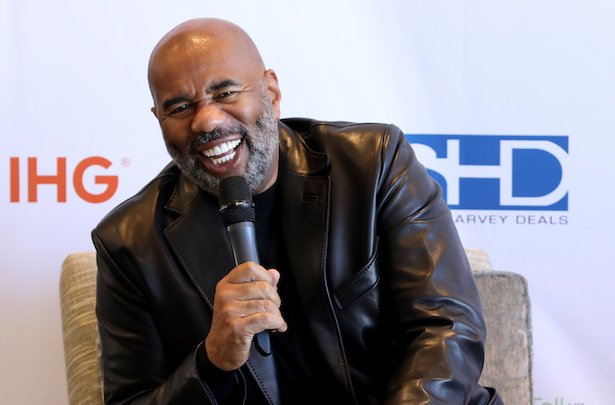 Steve Harvey in a leather jacket laughs against a white background