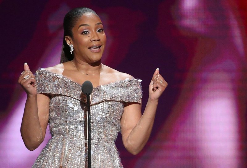 Tiffany Haddish talks into a microphone in a shiny silver dress with her hair in a ponytail