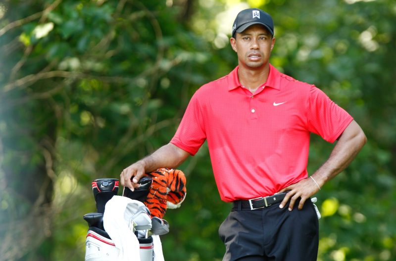 Tiger Woods in a bright red Nike polo resting one arm on his hip and the other on his clubs with a serious expression on his face.