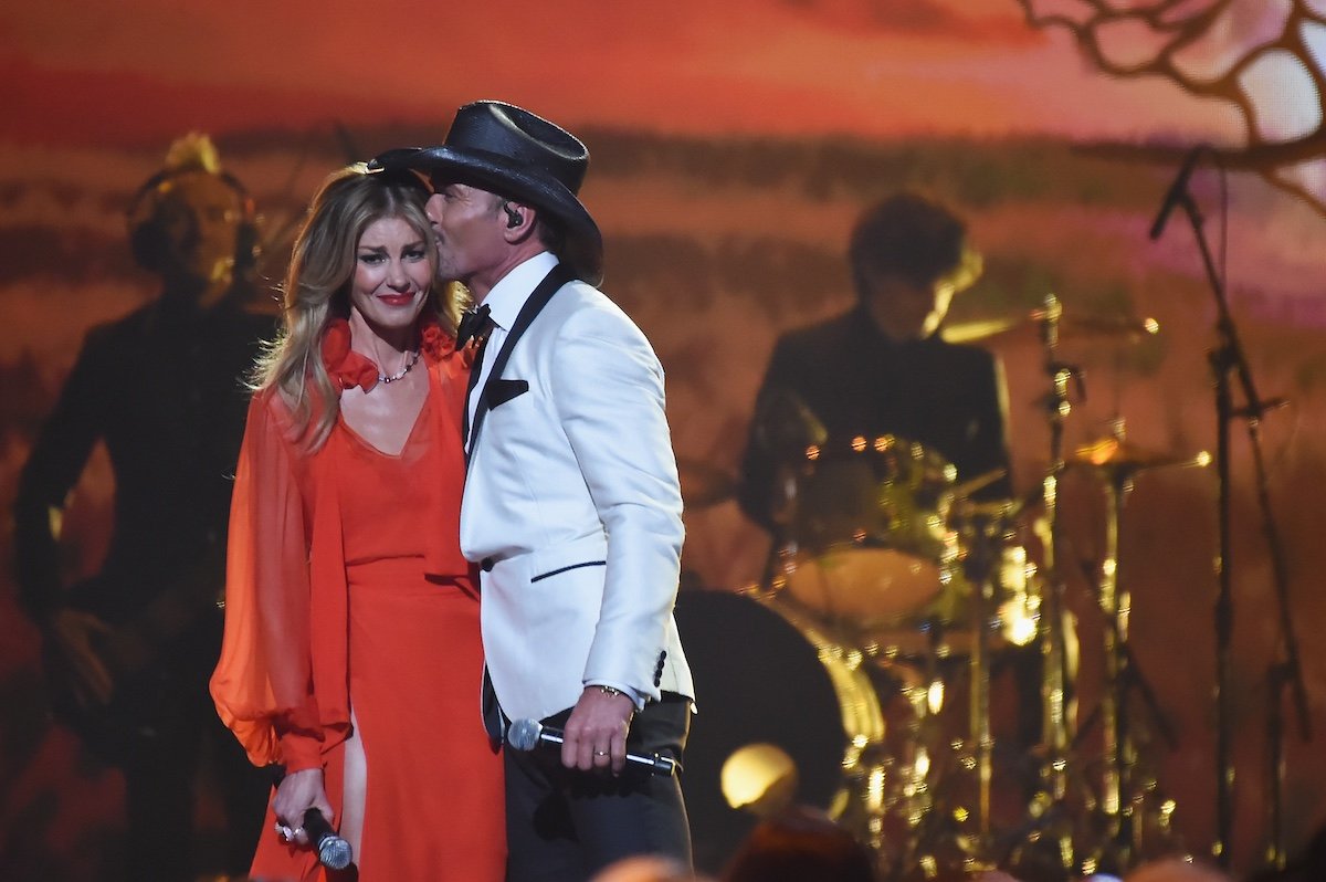 Tim McGraw in a white tuxedo and black cowboy hat kisses Faith Hill (in a red dress) on the side of