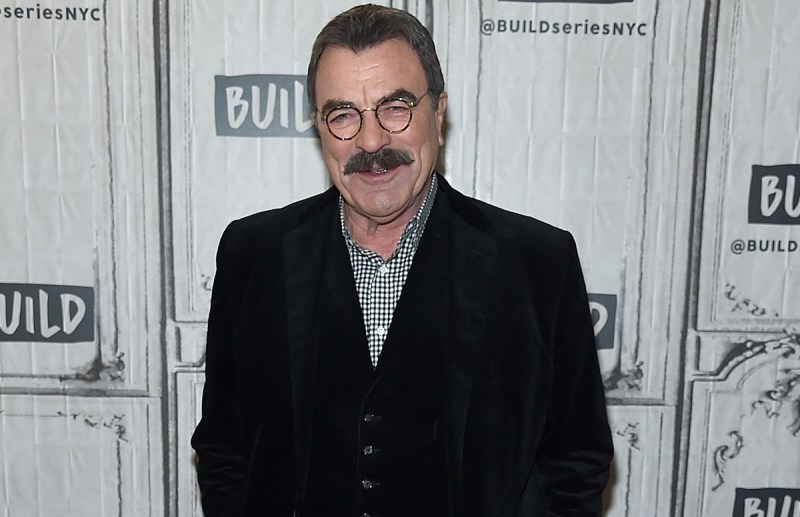 Tom Selleck smiling in a dark jacket and checkered shirt.