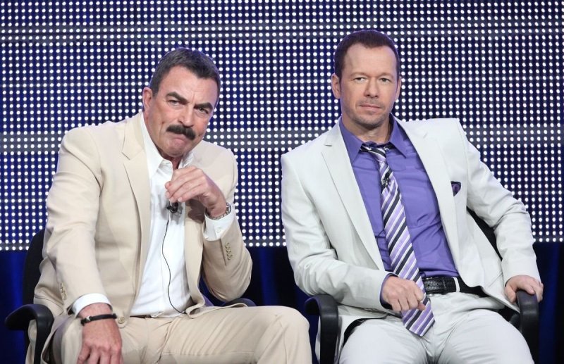 Tom Selleck wearing a beige suit sitting with Donnie Wahlberg, wearing a white suit, during a Blue B