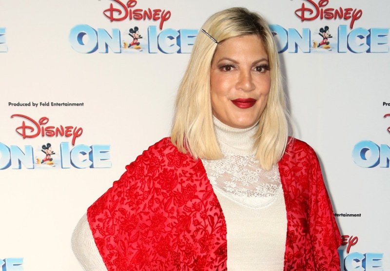 Tori Spelling smiles at the camera in a white top and red jacket against a white background