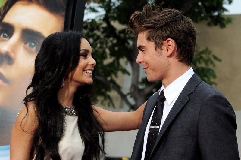 Vanessa Hudgens in a tan dress smiles and rests her arm on a smiling Zac Efron in a grey suit