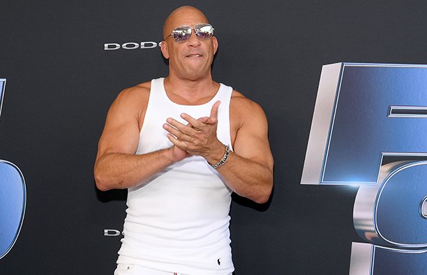 Vin Diesel in a white tank top, white jeans and sunglasses, clapping.