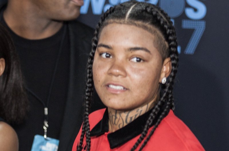 Young M.A. wears a red shirt and looks to the side as she stands in a group of people