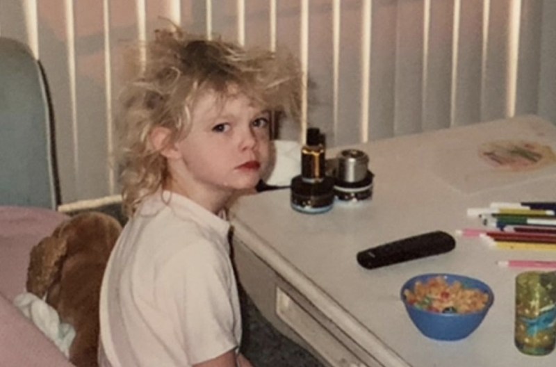 A small, blonde child wears a white tee shirt and looks grumpily at the camera in a living room
