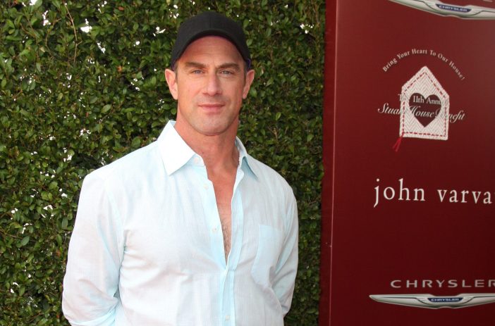 Christopher Meloni, dressed in a pale blue button up shirt and dark baseball hat, stands for photos