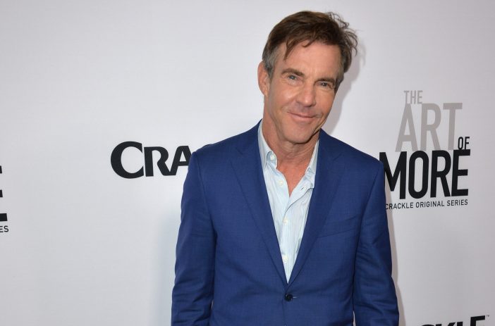 Dennis Quaid wears a blue suit while standing in front of a white background with black lettering