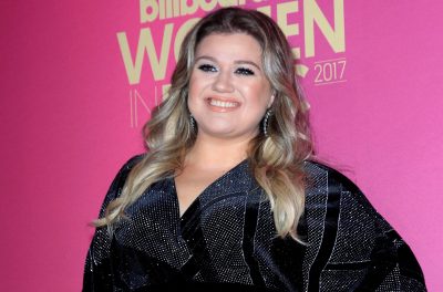 Kelly Clarkson wears a black dress covered in sparkles against a bright pink background