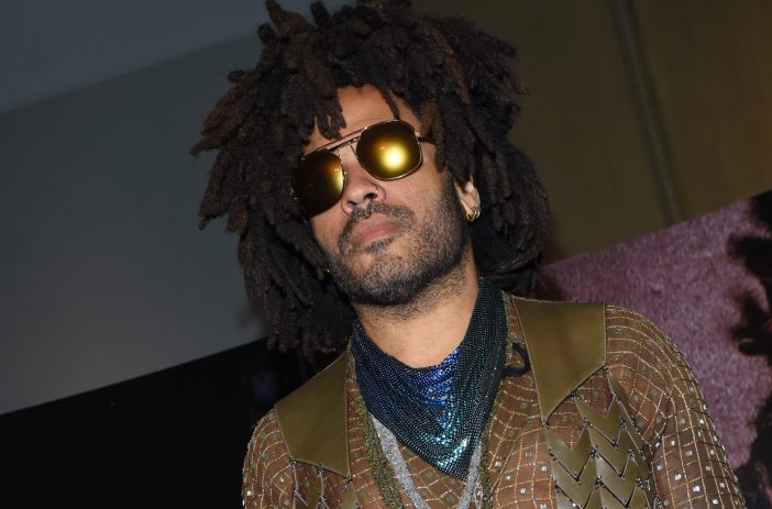 Lenny Kravitz wears a sheer shirt under a brown vest and blue scarf