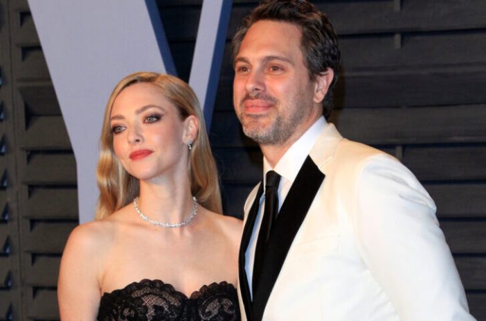 Amanda Seyfried and her husband Thomas Sadoski, posing on a red carpet. Amanda is wearing a black dress and Thomas is wearing a white suit with a black tie.
