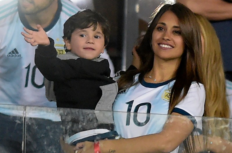 Antonella Roccuzzo wearing a blue and white jersey, holding a waving baby, and watching a soccer match.