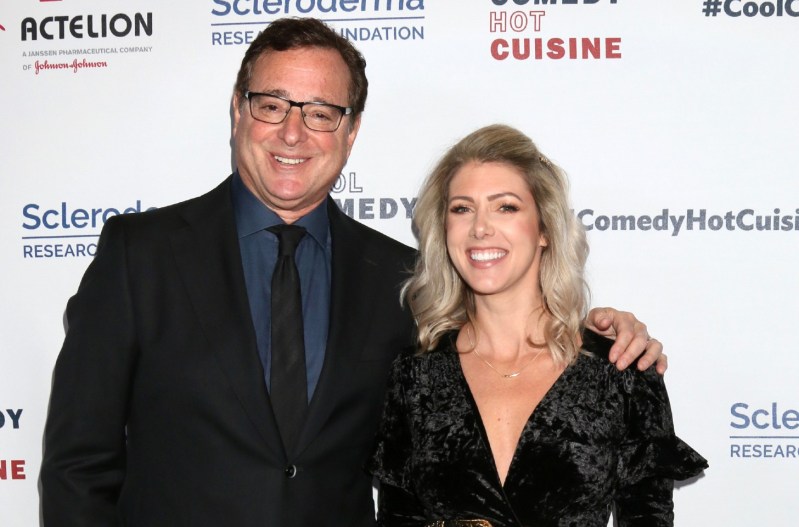 Bob Saget is wearing a black suit and smiling with his arm around his wife, Kelly Rizzo, who is smiling and wearing a black dress.