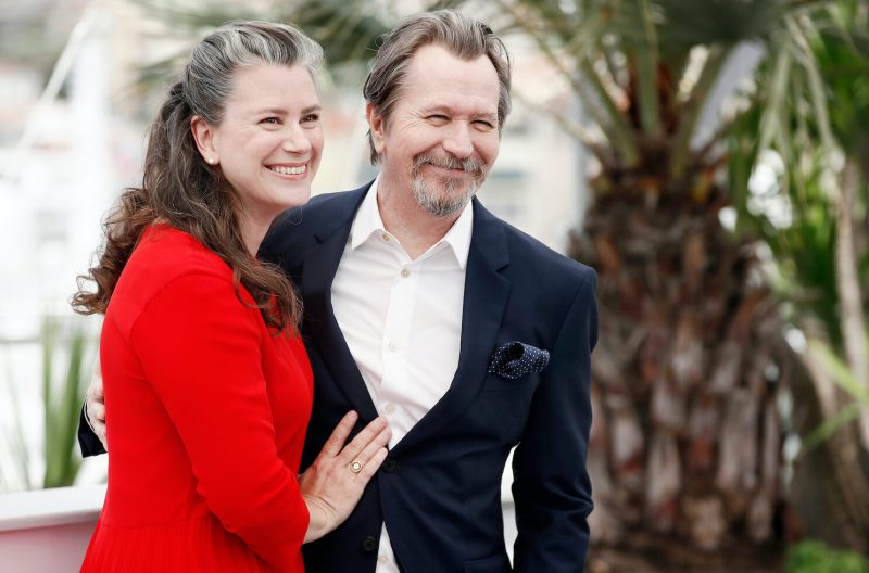 Actor Gary Oldman with his wife, Gisele Schmidt, on a red carpet. Schmidt is smiling and wearing a red dress with her hand rested on Oldman, who is wearing a black suit.
