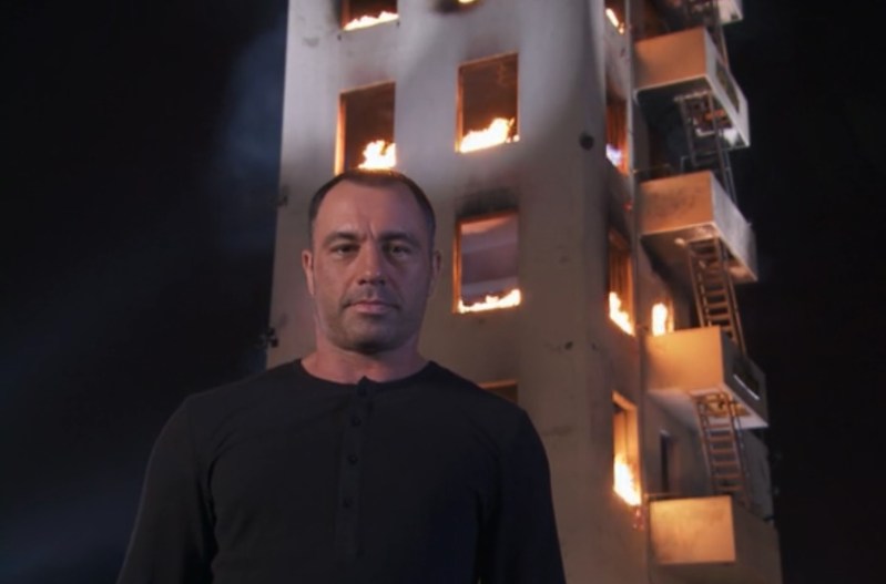 Joe Rogan standing in front of a burning building, wearing a black shirt and a blank facial expression.