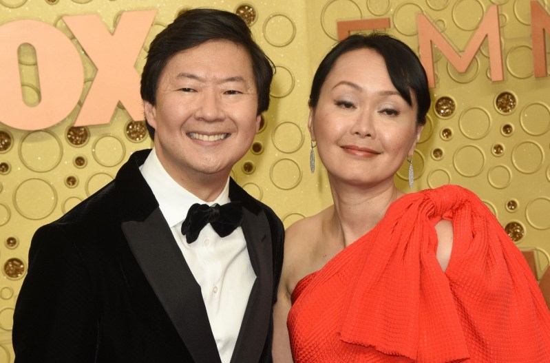 Ken Jeong is smiling and wearing a suit, standing with his wife, Tran Jeong, who is wearing an orange dress.