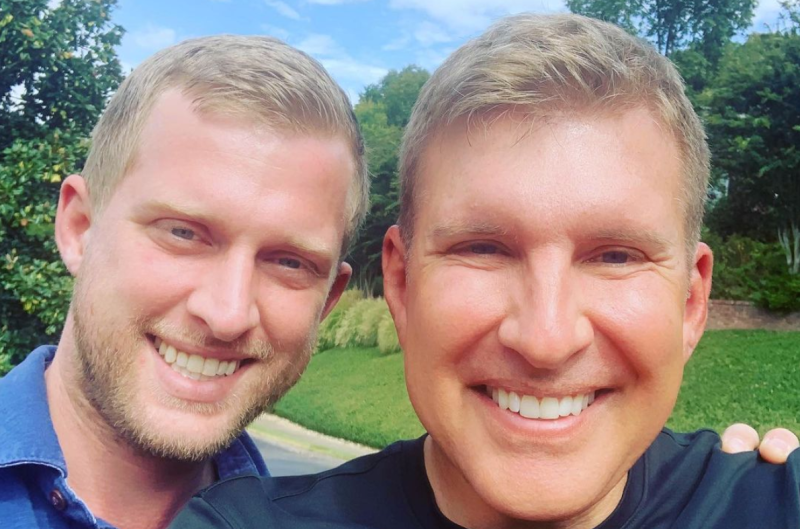 Todd Chrisley and his son, Kyle Chrisley, are standing outdoors wearing blue shirts and smiling.