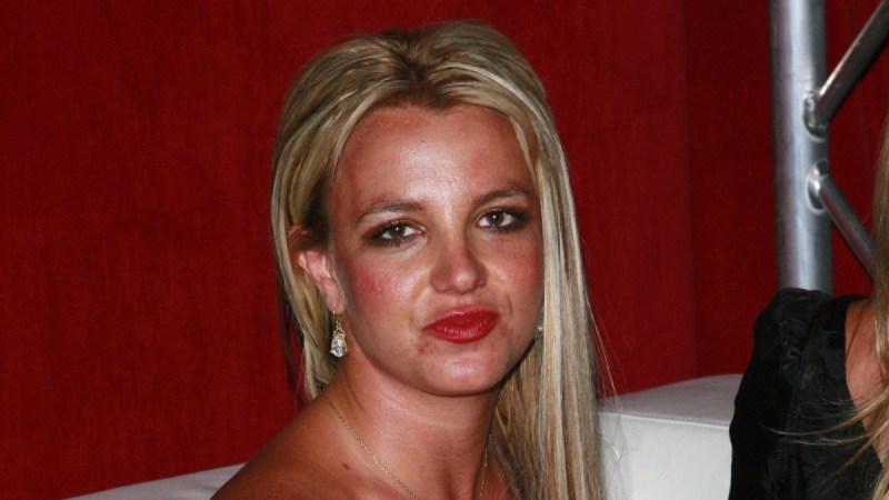 Britney Spears is seated on a white couch while wearing a black strapless dress