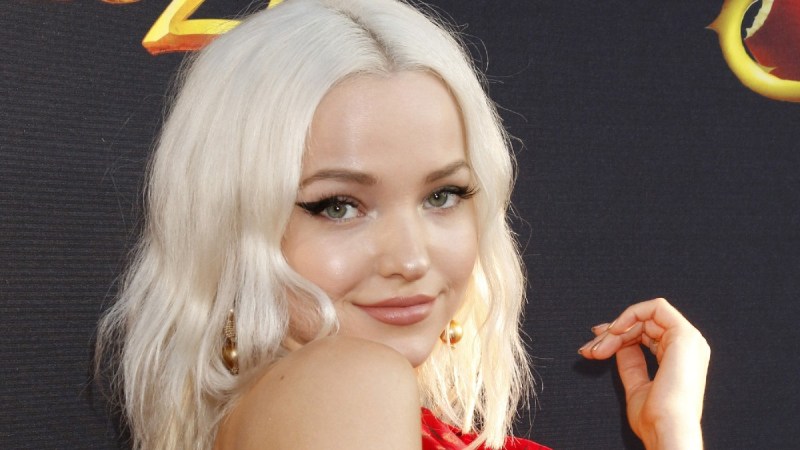 Dove Cameron, wearing a red dress, looks over her shoulder as she stands against a black background