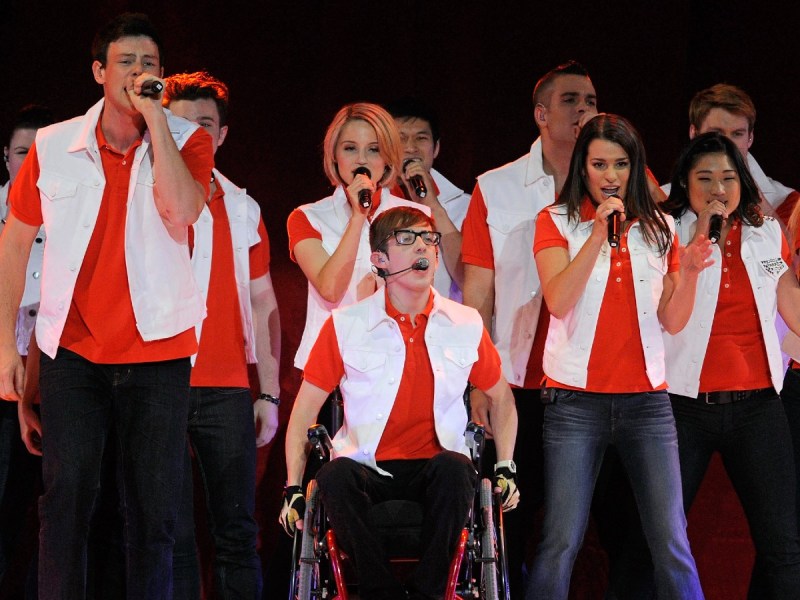 Glee cast dressed in matching red shirts with white vests singing into microphones onstage