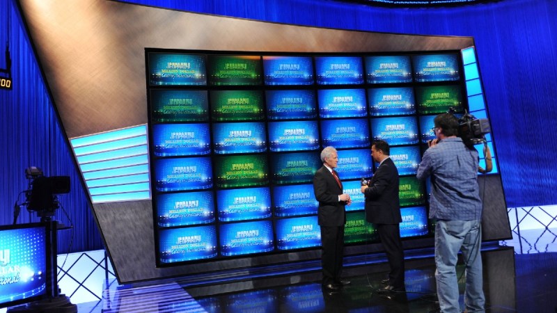 Alex Trebek speaks with another man on the set of Jeopardy