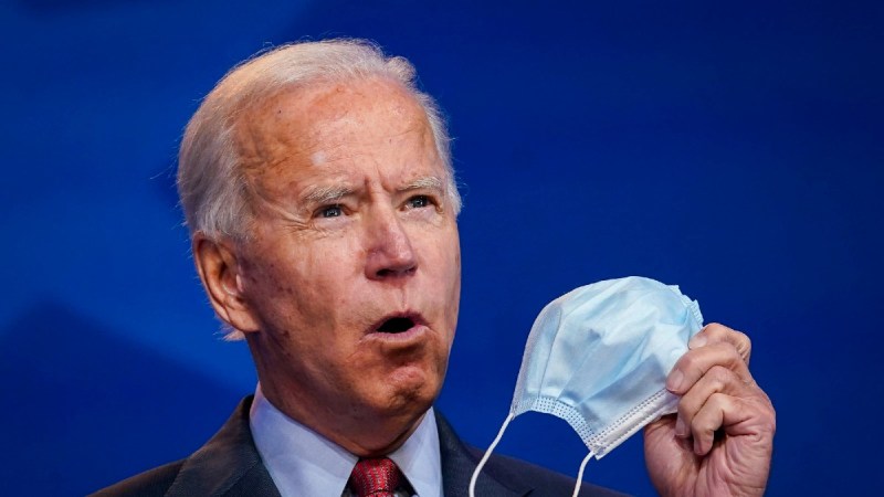 Joe Biden holds a pale blue face mask as he stands before a blue background
