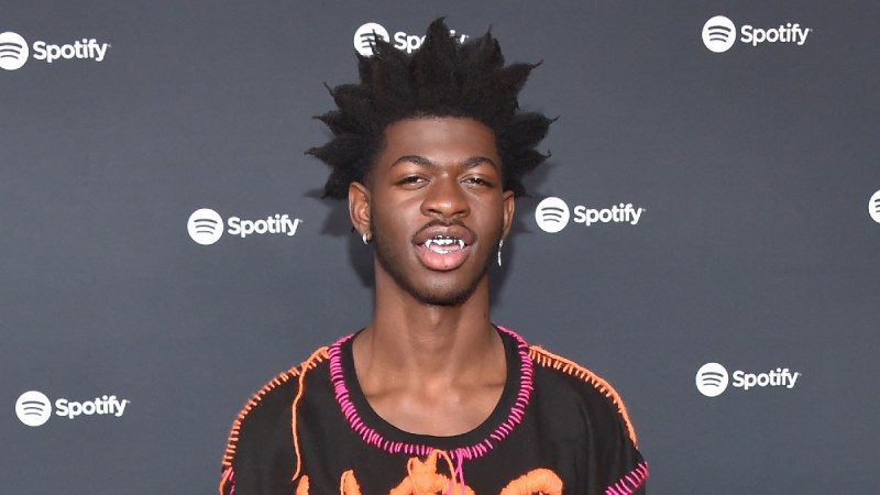 Lil Nas X wears a black shirt and silver grill against a black background