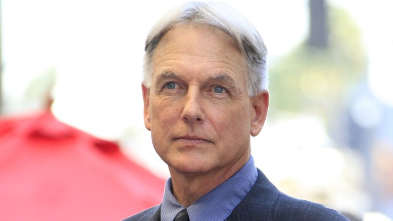 Mark Harmon wears a dark blue suit and stands in front of a red umbrella