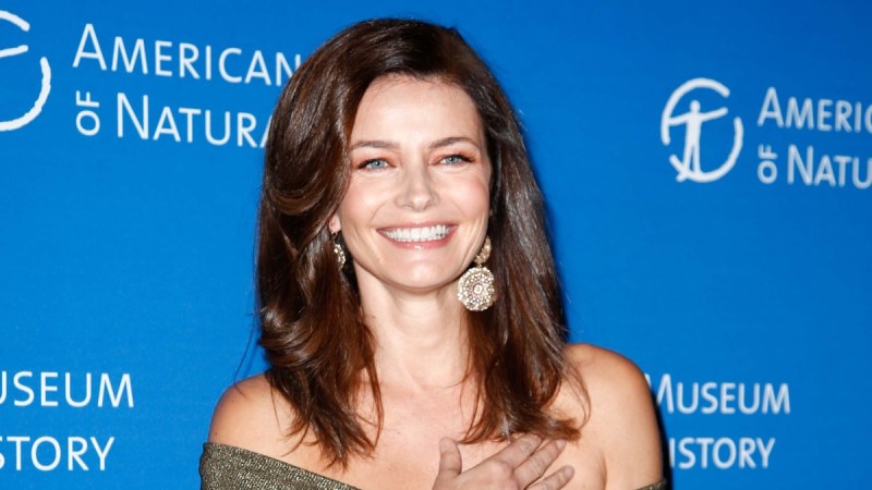 Paulina Porizkova wears an off the shoulder green dress in front of a blue background