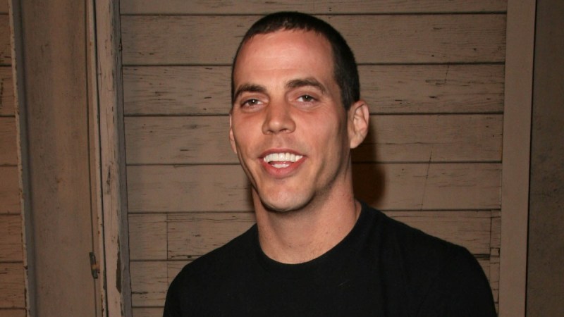 Steve-O wears a black t-shirt against a wooden background