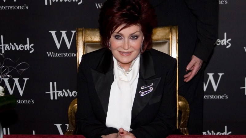 Sharon Osbourne wears a black suit jacket and white shirt as she sits in a golden chair at a red table