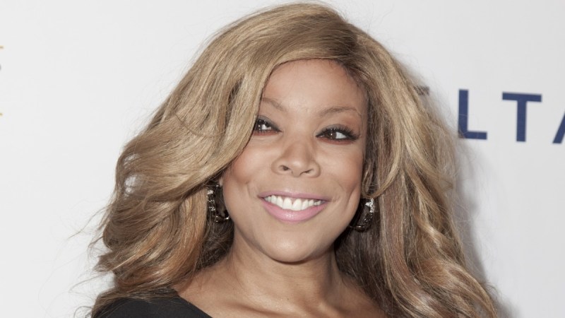 Wendy Williams smiles while wearing a dark colored dress against a white background