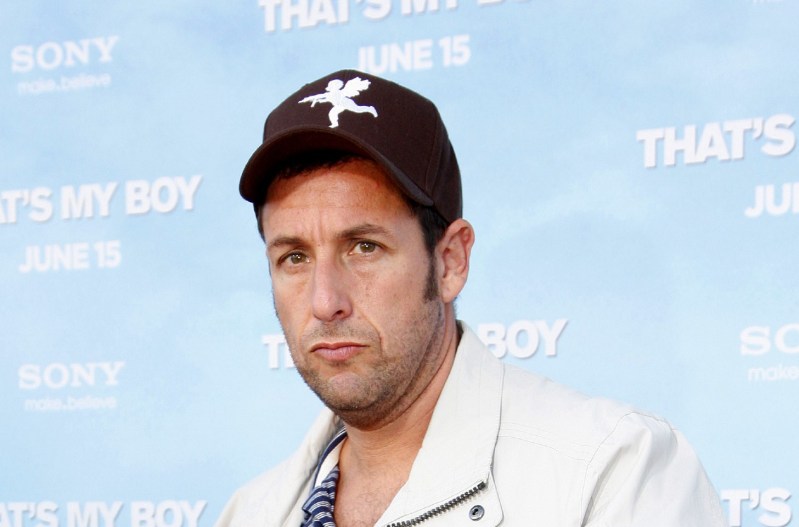 Adam Sandler pouting his lips and wearing a white jacket with a black baseball cap.