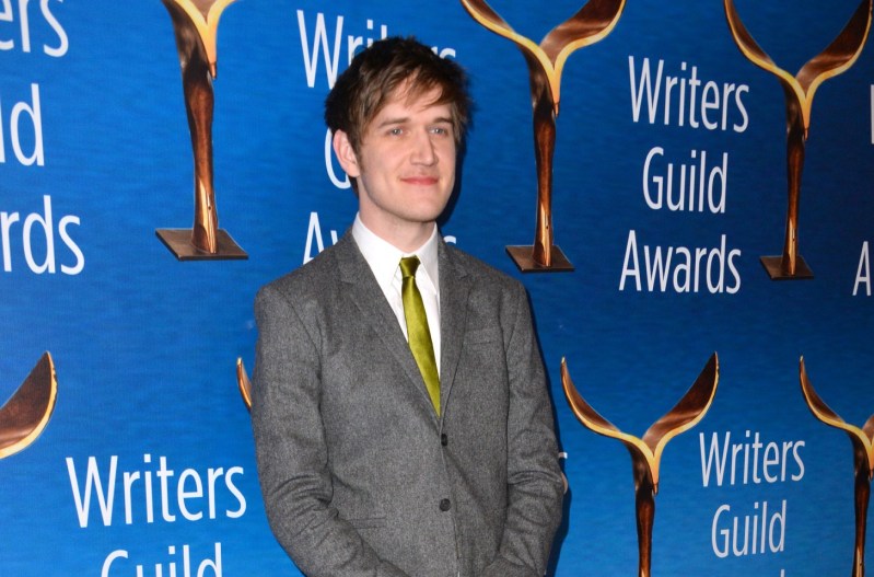 Bo Burnham smiling at the camera and wearing a grey suit with a green tie.