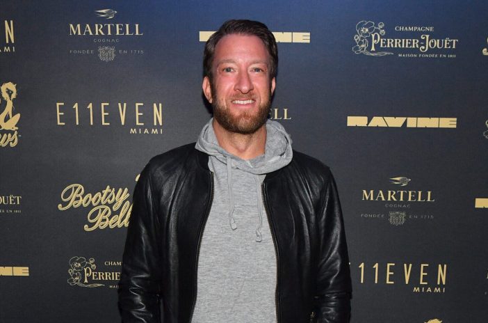 Dave Portnoy smiling in front of a step and repeat and wearing a black jacket and a grey hoodie.