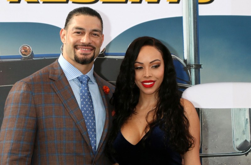 Roman Reigns in a grey suit with a blue shirt and tie, standing with his wife, Galina Becker, who is wearing a black dress.