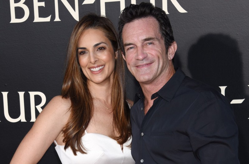 Jeff Probst and wife, Lisa Ann Russell, holding each other and smiling on the red carpet. Jeff is wearing a black shirt and Lisa is wearing a white dress.