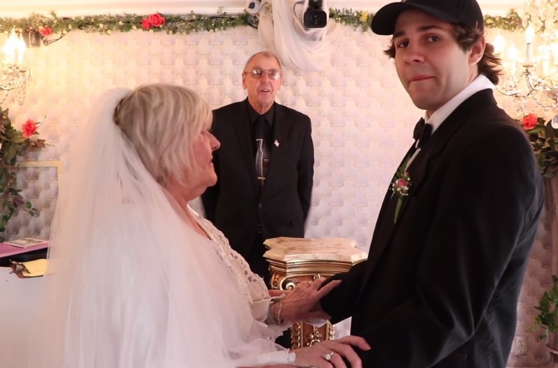 Lorraine Nash in a wedding dress holding hands with David Dobrik, who is wearing a black suit and a black baseball cap.