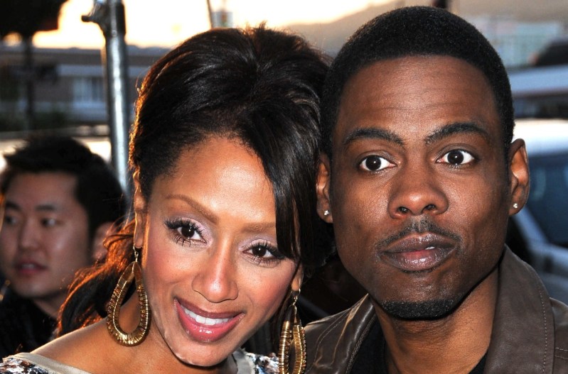 Chris Rock and his ex-wife Malaak Compton-Rock smiling at the camera. She is wearing a silver dress and gold earrings, and he is wearing a brown jacket.